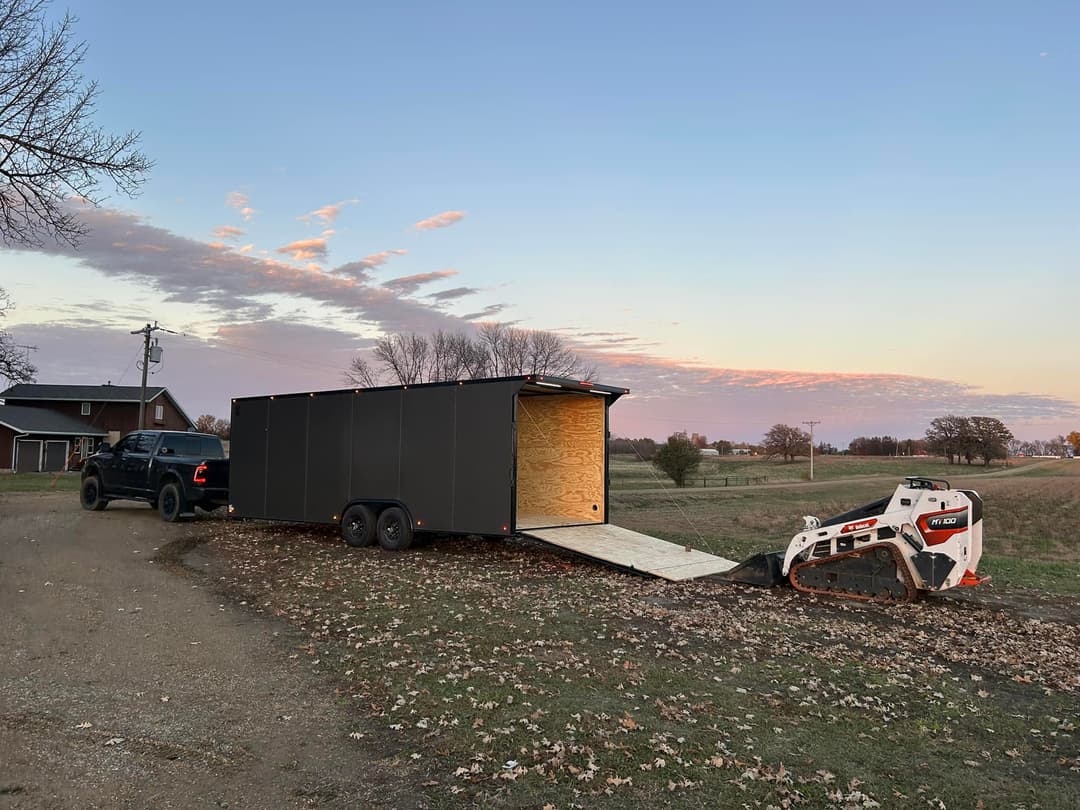 Truck towing a black trailer with loading ramp and skid steer loader at sunset in rural landscape.